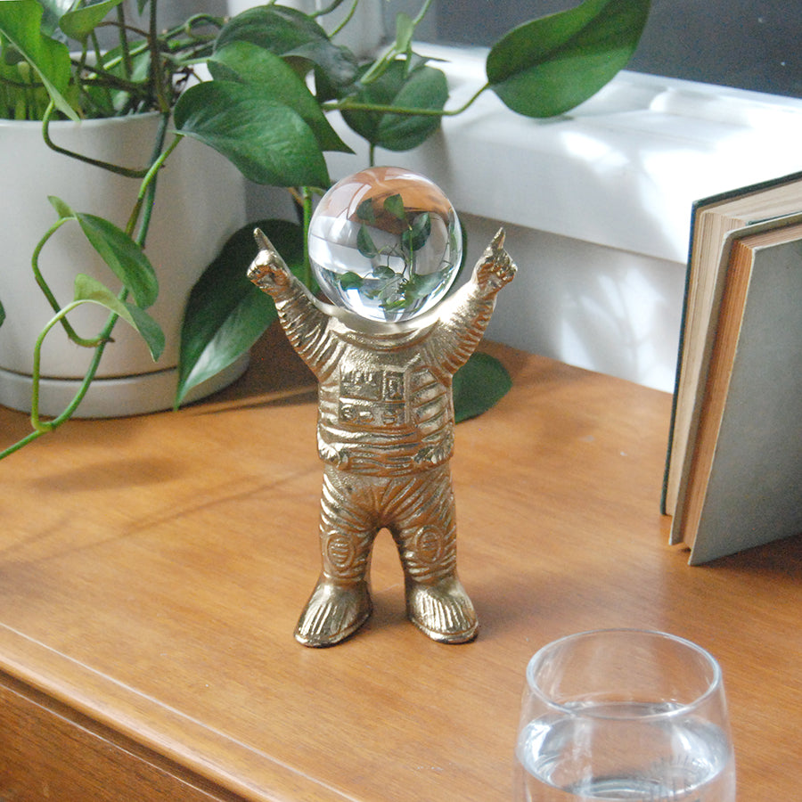 Gold astronaut figurine with a glass ball on a wooden surface with plants in the background