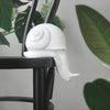 White resin snail sculpture perched on the edge of a black chair surrounded by greenery