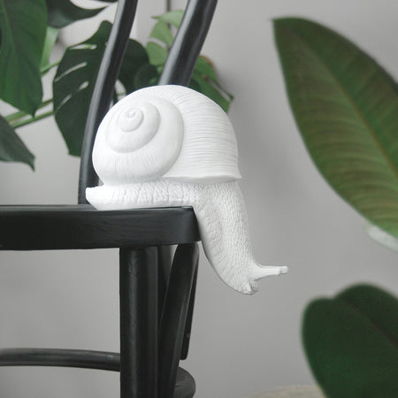 White resin snail sculpture perched on the edge of a black chair surrounded by greenery
